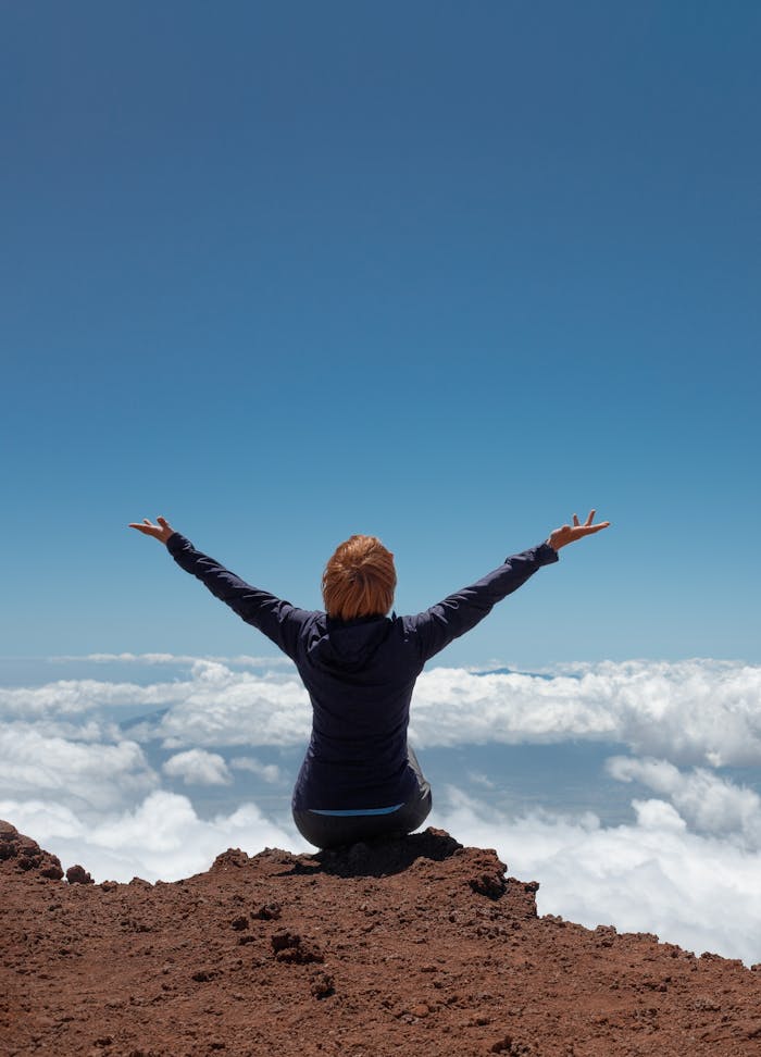 A woman sits atop a mountain, arms raised in triumph, overlooking clouds.
